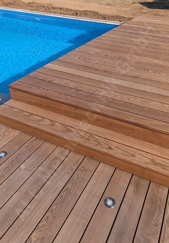 Poolrand mit Holzdeck-Stufe aus Thermoesche, Bodeneinbauleuchten und blauem Poolwasser