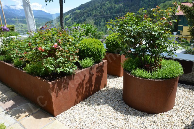 Eckige und runde Pflanzk&uuml;bel aus Cortenstahl auf einer Kiesfl&auml;che mit Bergblick