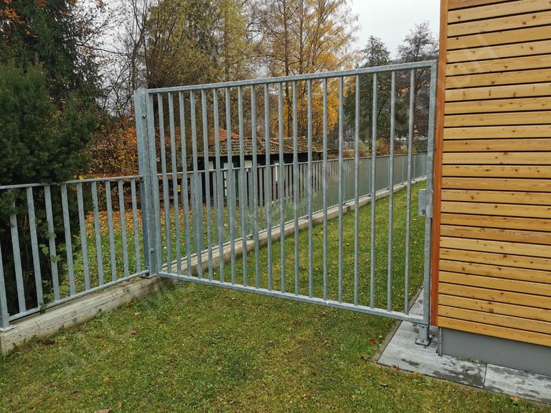 Verzinktes Gartentor aus Stabgitter mit passendem Stabzaun auf Betonsockel neben einer Holzfassade